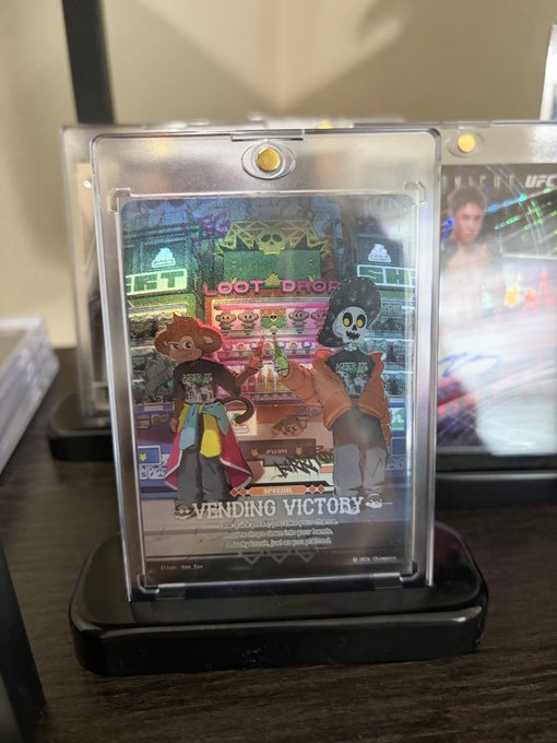 A holographic trading card in a protective display case featuring cartoon characters at a 'LOOT DROP' machine, with 'VENDING VICTORY' text at the bottom and what appears to be a UFC card visible behind it.