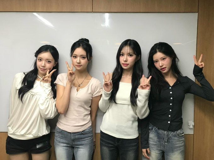 Four young women standing side by side in front of a whiteboard, each making a peace sign, dressed in casual tops and jeans.