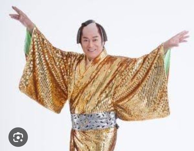 A person wearing a traditional Japanese gold kimono with an elaborate hairstyle stands with arms raised in a welcoming pose against a white background.