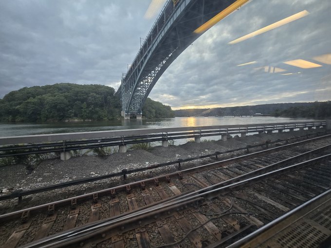 A view from a moving train shows railroad tracks, a large steel arch bridge spanning a river, and forested hills under a cloudy sky at dusk.