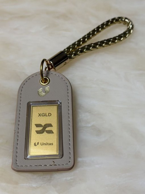 A physical gold bar labeled 'XGLD' and 'Unitas' encased in a grey leather keychain tag attached to a gold chain, resting on a light marble-like surface.