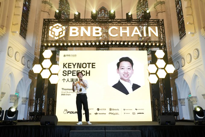 A man in a white shirt delivers a keynote speech on a large stage with an ornate architectural setting, standing in front of a screen displaying "BNB CHAIN KEYNOTE SPEECH" and identifying the speaker as Thomasi, Chief Business at BNB Chain.