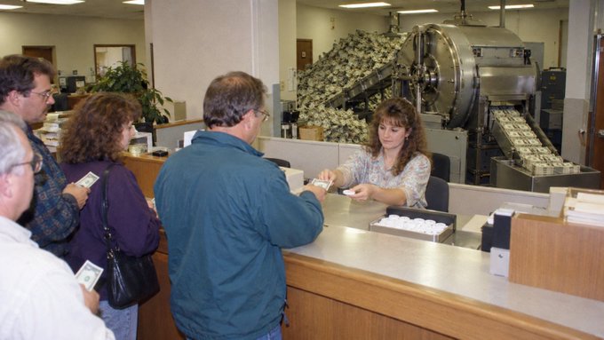 Customers wait in line at a bank teller window while a female employee assists them, with a large money-sorting machine visible in the background processing stacks of currency.