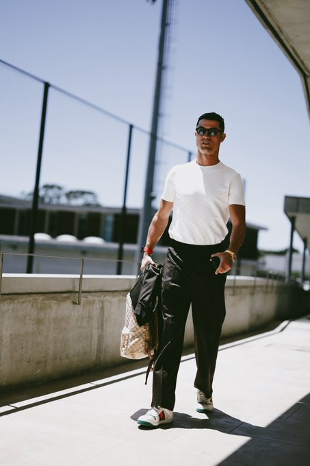 Street style photo of a man in a white t-shirt and black pants carrying a backpack while walking outdoors near a fenced area.