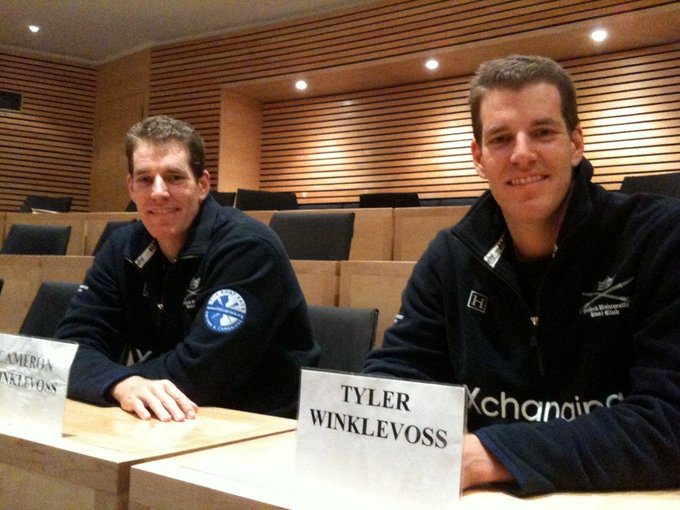 Tyler and Cameron Winklevoss sit at a panel with name plates identifying "TYLER WINKLEVOSS" and part of "CAMERON WINKLEVOSS" visible, wearing matching dark jackets as they smile at the camera in an auditorium-style setting. The image connects to the update highlighting their estimated $1.7 billion Bitcoin gains since 2015 and their continued $300M+ BTC holdings.