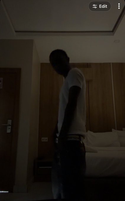 A dimly lit hotel room showing a man in a white t-shirt standing in silhouette, with a bed and wooden headboard visible behind him and an 'Edit' button in the corner.