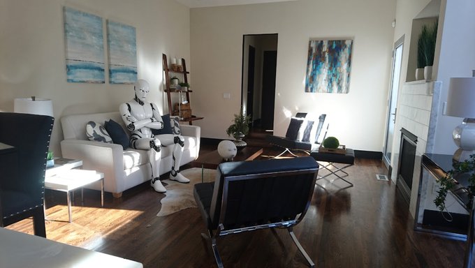 A modern living room with hardwood floors features a white couch, abstract blue artwork on the walls, a black lounge chair, and a humanoid robot sitting on the sofa next to throw pillows.