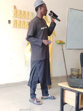 A man wearing traditional clothing and a knit cap speaks into a microphone while standing in front of a whiteboard in a classroom-like setting.