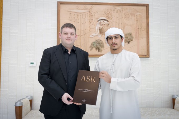 The image features two men standing next to each other with one man holding a book titled "ask" while the other man holds a sign that says "ask". The background includes a painting of a mosque, which suggests that they are in an Arabic-speaking country or region. The text on the post indicates that there is a joint venture between Keeta and ASK Group, led by His Highness Sheikh Ahmed bin Sultan bin Khalifa bin Zayed Al Nahyan, aiming to tokenize tens of billions of dollars of commodities and modernize cross-border transactions in the Middle East.