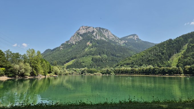 A scenic landscape photograph of a calm lake with greenish water reflecting a large rocky mountain surrounded by dense forest under a clear blue sky.