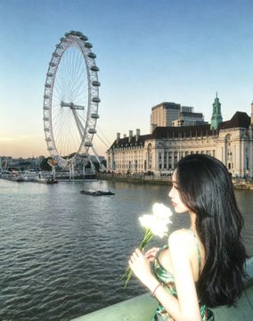 A woman with long dark hair holds a white rose in the foreground, with the London Eye and County Hall visible across the River Thames at sunset.