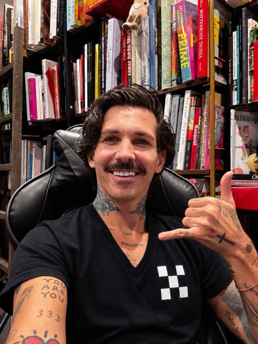 A smiling man with a mustache and tattoos sitting in front of a large bookshelf filled with books, including visible titles like "FRUiTS" and works by Weston Noguchi Tanaka.
