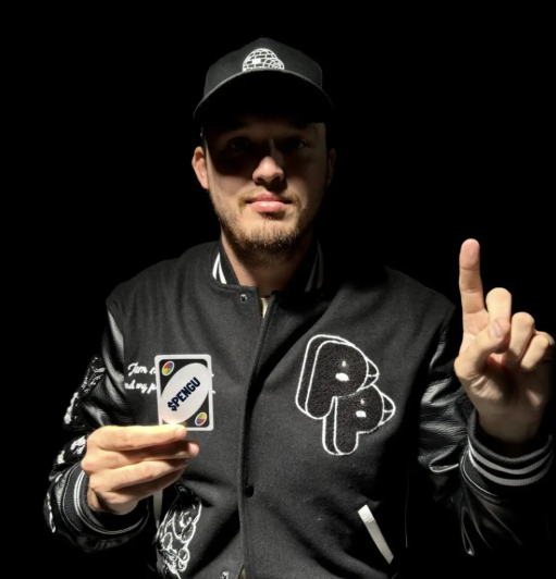 A man wearing a black cap and a black 'P' varsity jacket holds up a card reading 'SPENGLI' while gesturing with his other hand against a black background.