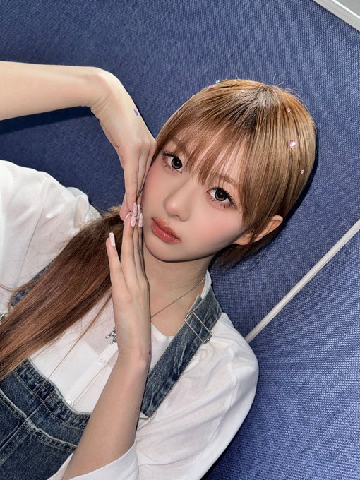 A young woman with light brown hair and decorative hair clips poses for a selfie, wearing a white blouse and denim overalls, lying on a blue textured surface.