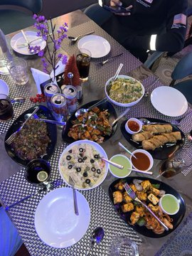 A spread of various Asian dishes—sushi, dumplings, stir-fry, spring rolls, and dipping sauces—is laid out on a dining table, suggesting a feast that appears to cost more than the $24 the poster claimed to have spent. The caption playfully questions whether the meal was a win or loss in value given the generous portions and variety shown.