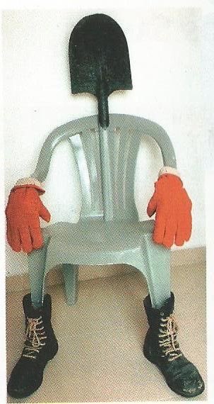 A surreal anthropomorphic sculpture features a gray plastic chair topped with a black shovel for a head, wearing orange work gloves as hands resting on its armrests, and black work boots on its legs—humorously embodying the "Chair is Chair" post by transforming the chair into a complete "lazy" figure unwilling to fulfill its seating purpose.