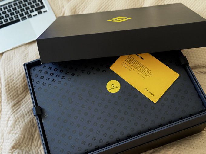 An open black box branded with a yellow Binance logo sits on a bed next to a laptop, containing a patterned insert and a yellow informational card.