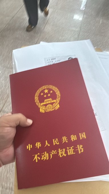 A hand holds a red Chinese real estate certificate titled '中华人民共和国不动产权证书' over a white envelope.