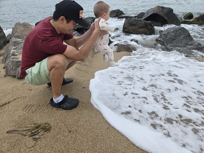 A man wearing a black cap with 'RF' on it, a maroon shirt, and green shorts crouches on a beach to hold a baby near the foamy edge of the ocean waves.