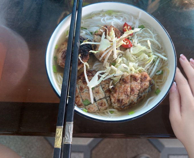 A bowl of Vietnamese-style noodle soup, likely bún chả, topped with grilled meat patties, shredded cabbage, and chili slices, with chopsticks resting across the rim and a hand visible on the side.