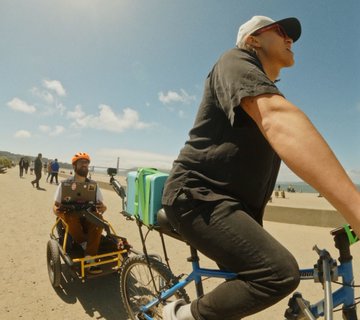 This image shows a man riding a bicycle, with another person pulling a wheelchair behind him. The wheelchairs' occupant appears to be a person wearing a white cap who is actively engaging with the motion graphics editor via text. The scene captures the essence of modern technology and human interaction by combining traditional activities (cycling) with contemporary digital experiences (motion graphics).