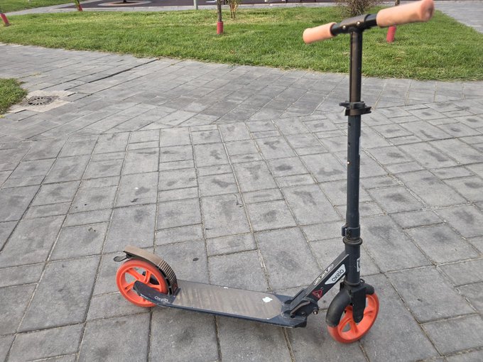 A black and orange Oxelo adult scooter with tan handle grips parked on a paved stone walkway near grass.