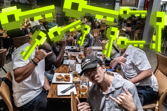 A group of men posing at a restaurant table with beer and food, making hand gestures, with green pixelated Japanese text overlaid on the image.
