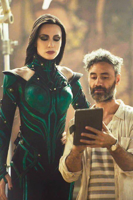 Two actors in costume on a film set - a woman in an elaborate teal and black armor-like outfit and a bearded man in casual attire reading from a tablet, likely a behind-the-scenes shot.