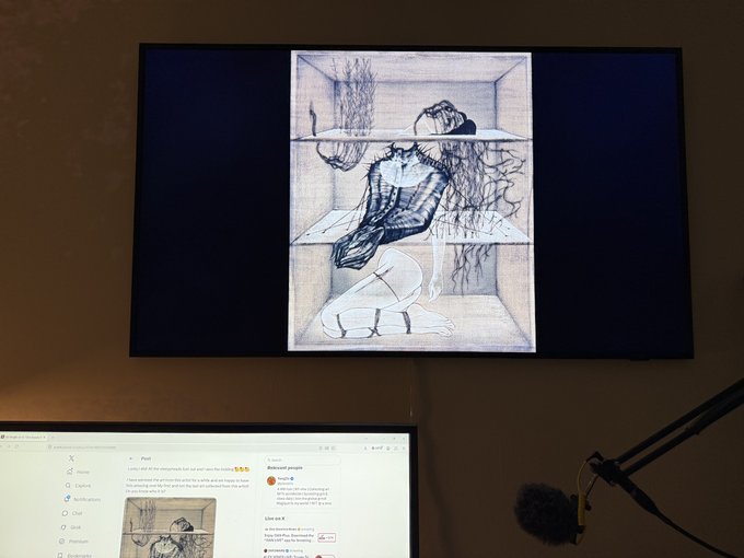 A flat-screen TV mounted on a wall displaying a sketch of a kneeling figure, with a computer monitor showing a Reddit post below and a microphone on a stand to the right.