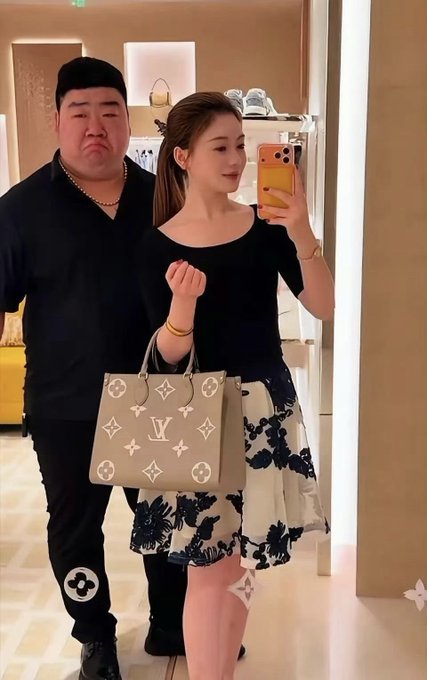 A man in black attire and a woman in a black top with floral skirt take a mirror selfie, with the woman holding a Louis Vuitton handbag.