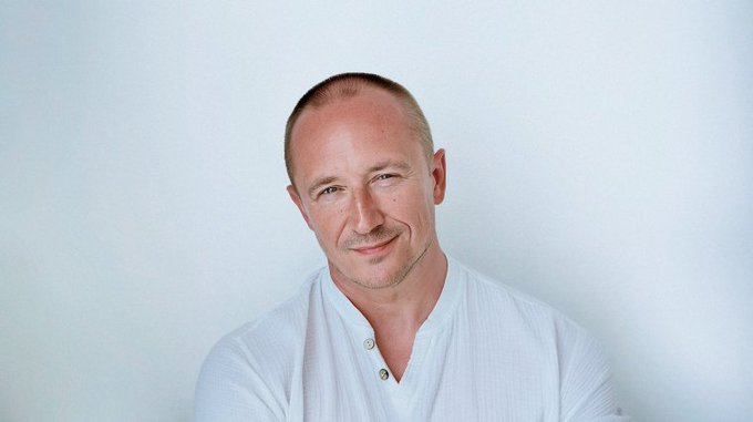 A portrait of a bald man with light stubble wearing a white henley shirt, smiling slightly against a plain light blue background.