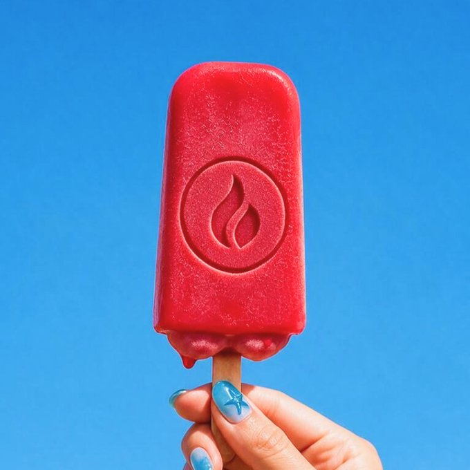 A hand with blue star-patterned nail polish holding up a red popsicle embossed with a flame logo against a clear blue sky.
