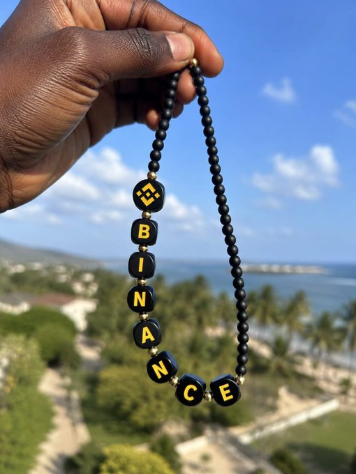 A hand holds up a beaded necklace spelling "BINANCE" with yellow letters on black cube beads, set against a scenic tropical ocean backdrop—fittingly paired with a cheerful "Gm!" greeting for the crypto community.