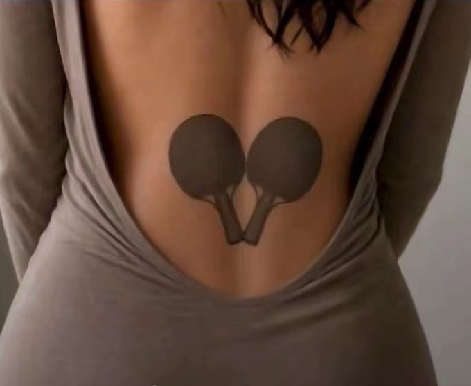 A back tattoo of two spoons with handles crossing at the bottom, resembling a heart shape between the shoulder blades of a person wearing a gray tank top.