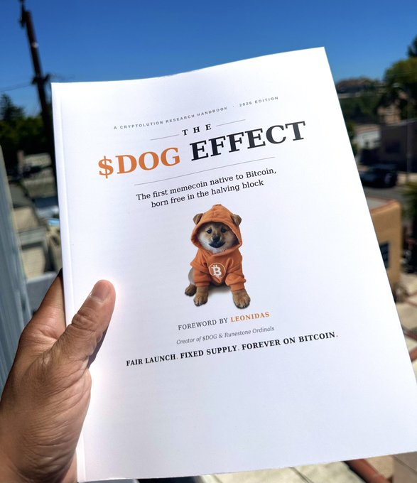 A hand holds up a white paperback titled "THE $DOG EFFECT — A Cryptolution Research Handbook, 2026 Edition," featuring a Shiba Inu in an orange Bitcoin-logo hoodie on the cover, with a foreword by Leonidas (creator of $DOG & Runestone Ordinals) and the tagline "Fair launch. Fixed supply. Forever on Bitcoin."