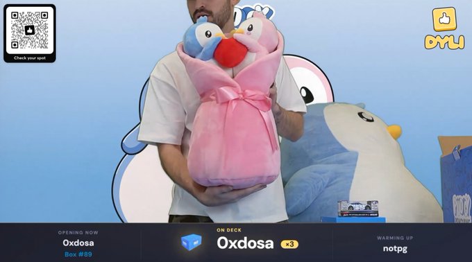 A streaming/giveaway overlay featuring a person holding a pink plush toy with two penguin-like characters, branded 'DYLI' in the top right, with boxes listed at the bottom: 'Oxdosa Box #89' and 'Oxdosa +3' warming up with 'notpg.'