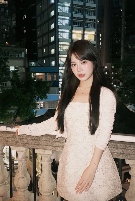 A woman with long dark hair, wearing a white textured mini dress and delicate gold jewelry, poses on an ornate balcony at night with illuminated city skyscrapers in the background. The image ties to the post as a stylish memento captured during her busy Hong Kong trip, where she attended the @BNBCHAINZH event before heading home.