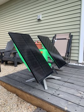 The image features a wooden deck with two solar panels mounted on it, both facing towards the sun. There are also two chairs placed on the deck, one of which has a green cushion. The background shows a house exterior, indicating that this outdoor space is located in someone's backyard or patio area.