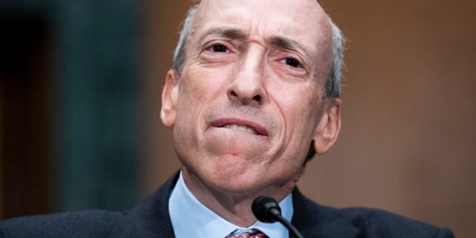 A close-up photograph of an older man in a suit and tie speaking into a microphone.