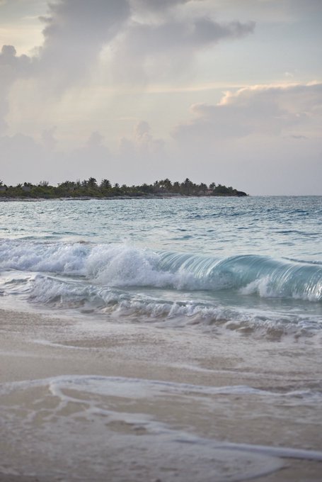 Gentle waves roll onto a sandy beach, with a tropical island covered in palm trees visible across the water under a cloudy sky.