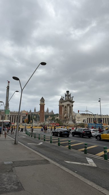 An overcast street view of Plaça d'Espanya in Barcelona, showing the Venetian towers, Montjuïc communications tower, and traffic.