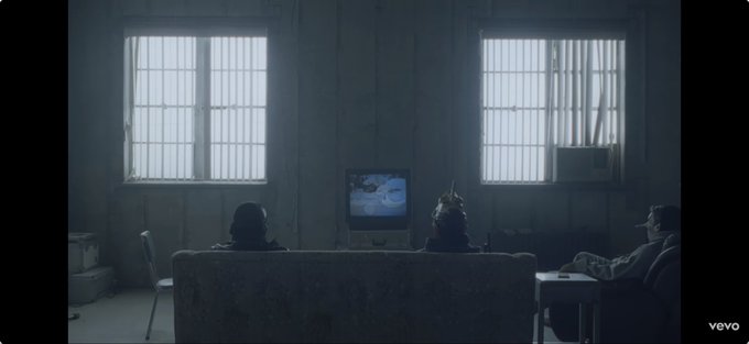 Two figures sit on a couch watching a grayscale TV in a dim, cold room, while a third person sits separately at a side table, visually echoing the bleak, isolated "deep winter" referenced in the post about Bitcoin miners' fee revenue hitting a 10-year low. The VEVO watermark appears in the bottom right corner of the frame.