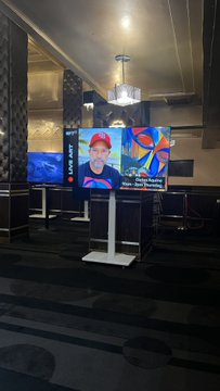 A dimly lit event space with a digital screen displaying a man's face wearing a red "MAKE AMERICA GREAT AGAIN" hat, alongside abstract artwork on a white stand.