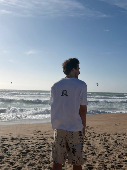 A man stands on a sandy beach facing the ocean under a bright blue sky, wearing a white t-shirt with a small black graphic and patterned shorts, with kitesurfers visible in the distance.