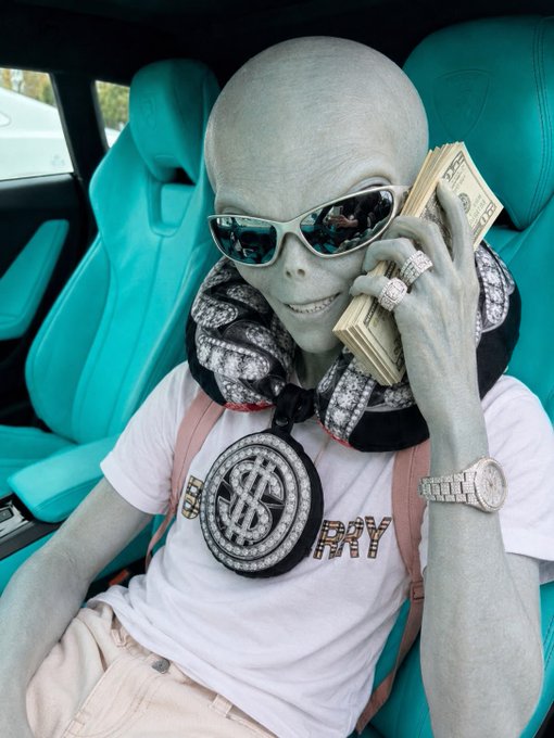 A gray-skinned alien figure wearing sunglasses, a large dollar-sign pendant necklace, and holding a stack of cash while sitting in a teal luxury car interior.