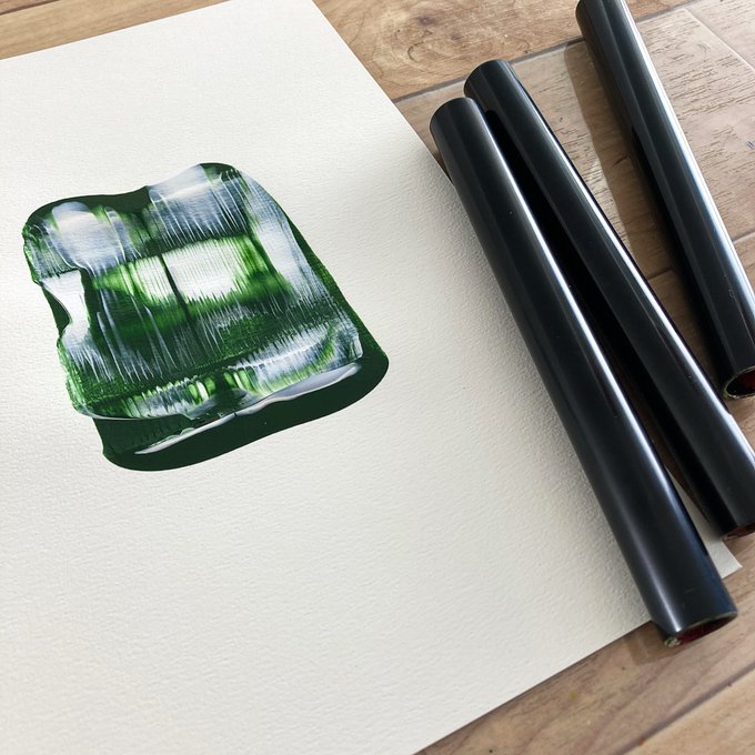A photograph of an abstract green and white painting on a textured paper canvas, placed next to several black rolled tubes on a wooden floor.
