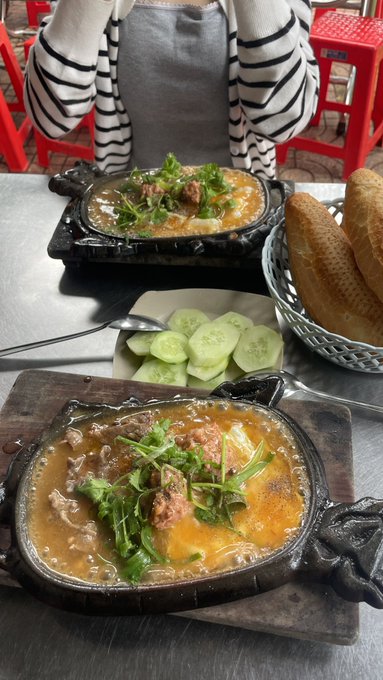 Two sizzling cast iron skillets of food topped with cheese and herbs are served alongside a basket of bread and a plate of sliced cucumbers.