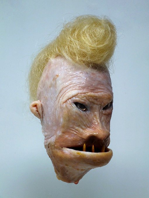 A grotesque, fleshy sculpted mask or doll head with blonde hair styled in a pompadour, featuring wrinkled pink skin, sunken eyes, and a wide mouth with sharp yellow teeth.
