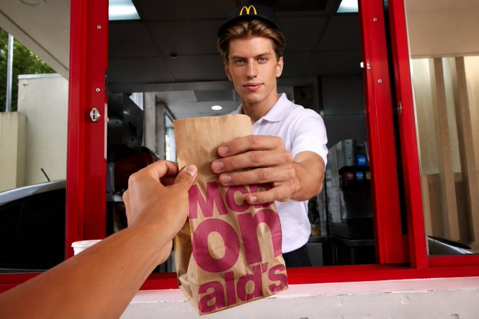 A young McDonald's employee wearing a branded cap hands a brown paper bag through a drive-thru window to a customer's outstretched hand.