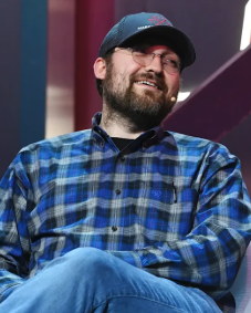 A bearded man in a blue plaid shirt and baseball cap smiles while looking off to the side, appearing to be Charles Hoskinson, the Cardano founder mentioned in the post announcing his development of a new programming language and plans involving Midnight ($NIGHT).
